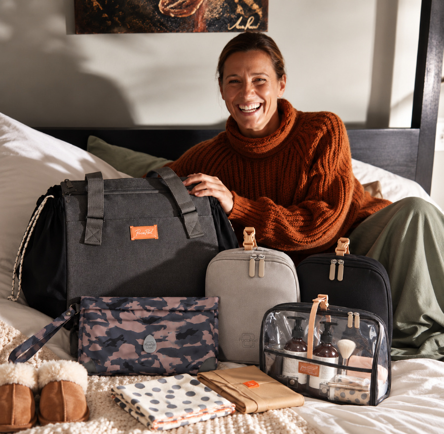 A smiling woman sits on a bed surrounded by multiple travel and toiletry bags from the brand Chloé. The collection includes a large dark gray duffel bag, a light gray zippered bag, a black bag, a clear zippered toiletry bag with bottles, a camouflage pouch, and folded fabric accessories on a beige knit blanket.