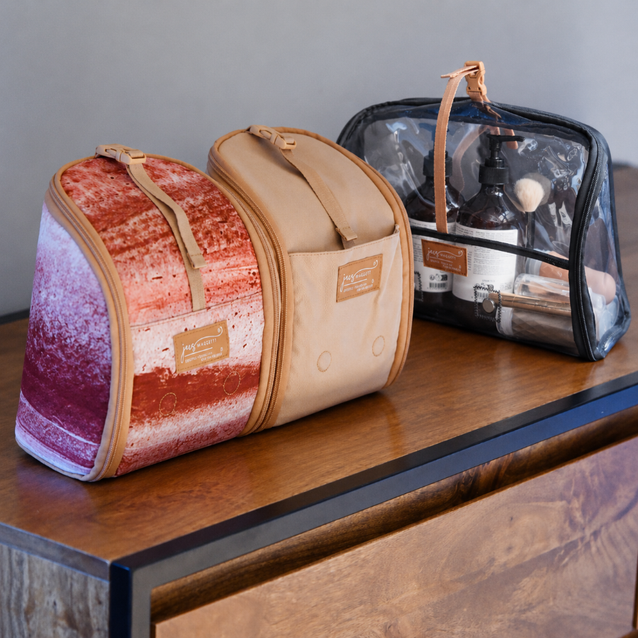 Three cosmetic or travel pouches displayed on a wooden table against a gray wall. The front pouch is beige with a front pocket and a tan leather patch.