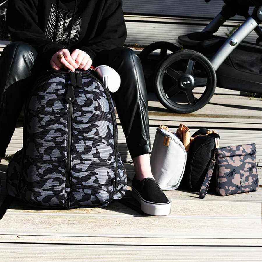 A person sitting outdoors on wooden steps next to a black and gray camouflage backpack, with a stroller wheel and other bags in the background.